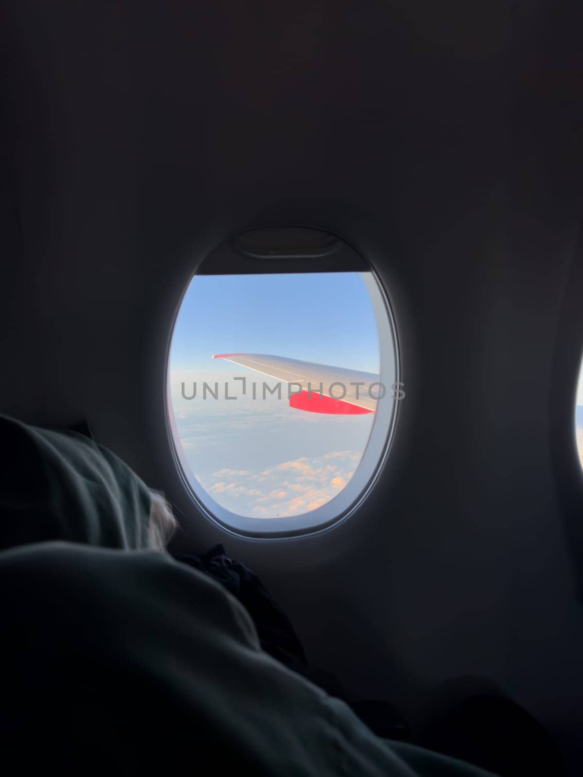View from airplane window showing wing above clouds during peaceful flight, with passenger resting in seat, soft light creating calm travel atmosphere