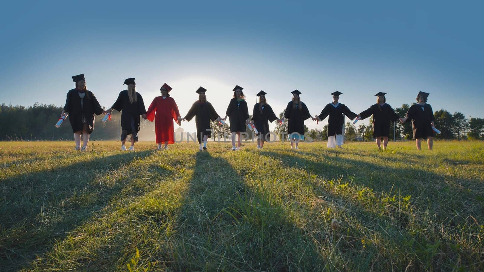 Group of graduates is walking in a field at sunset, holding hands and their diplomas