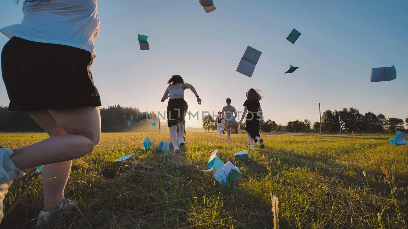 Group of high school students running through a field throwing their notebooks in the air celebrating the end of school at sunset.