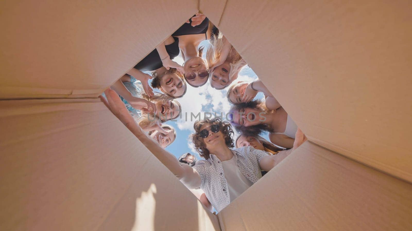 Group of surprised high school students looking inside a large cardboard box during summer, expressing excitement and curiosity