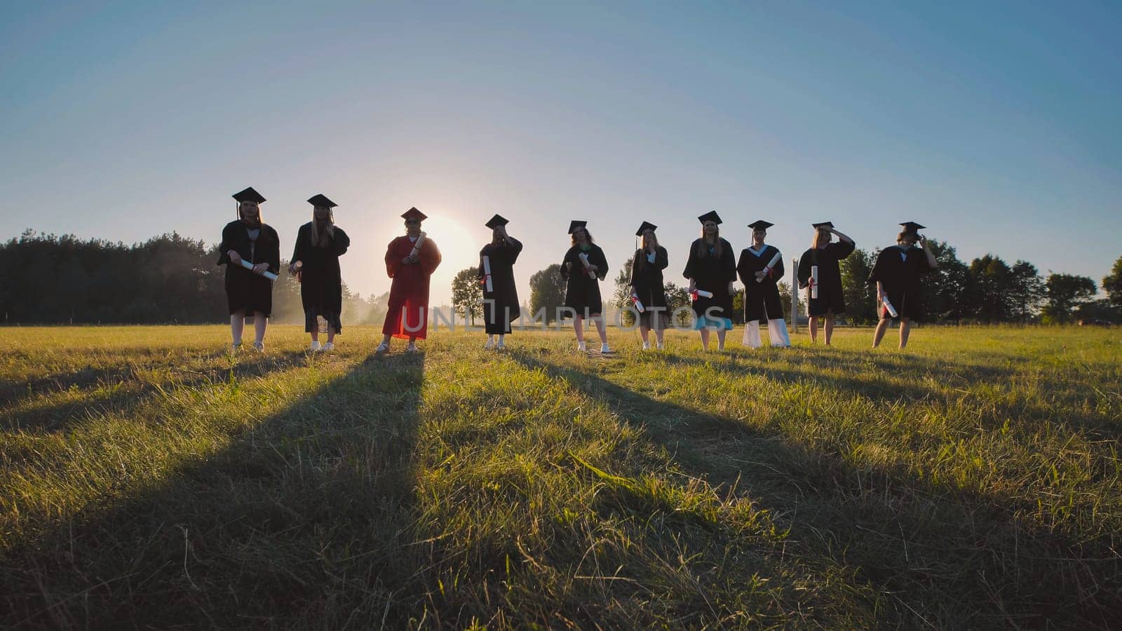 Group of graduates wearing academic gowns and holding diplomas, standing in a field at sunset, celebrating their academic achievement