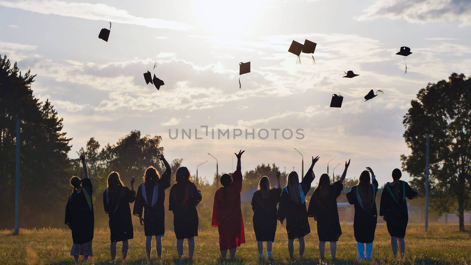 Group of joyful students in academic gowns celebrating graduation by throwing their caps high into the air against a stunning sunset backdrop