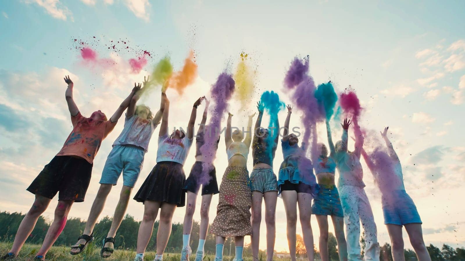 Group of cheerful school friends jumping and throwing colorful powder in the air against a sunset sky, celebrating friendship and creating vibrant memories