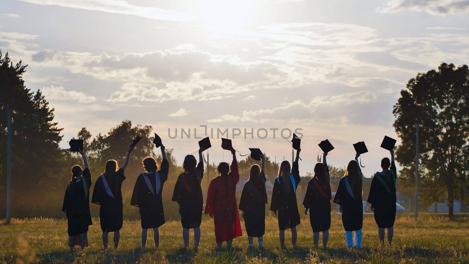 Group of graduates raising their mortarboards in a field at sunset, celebrating their academic achievement and the end of their studies