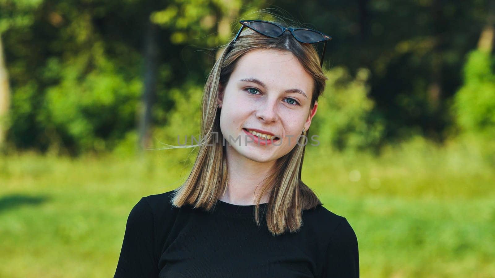 Portrait of a smiling blonde teenage student wearing a black t shirt and sunglasses on her head in a green park