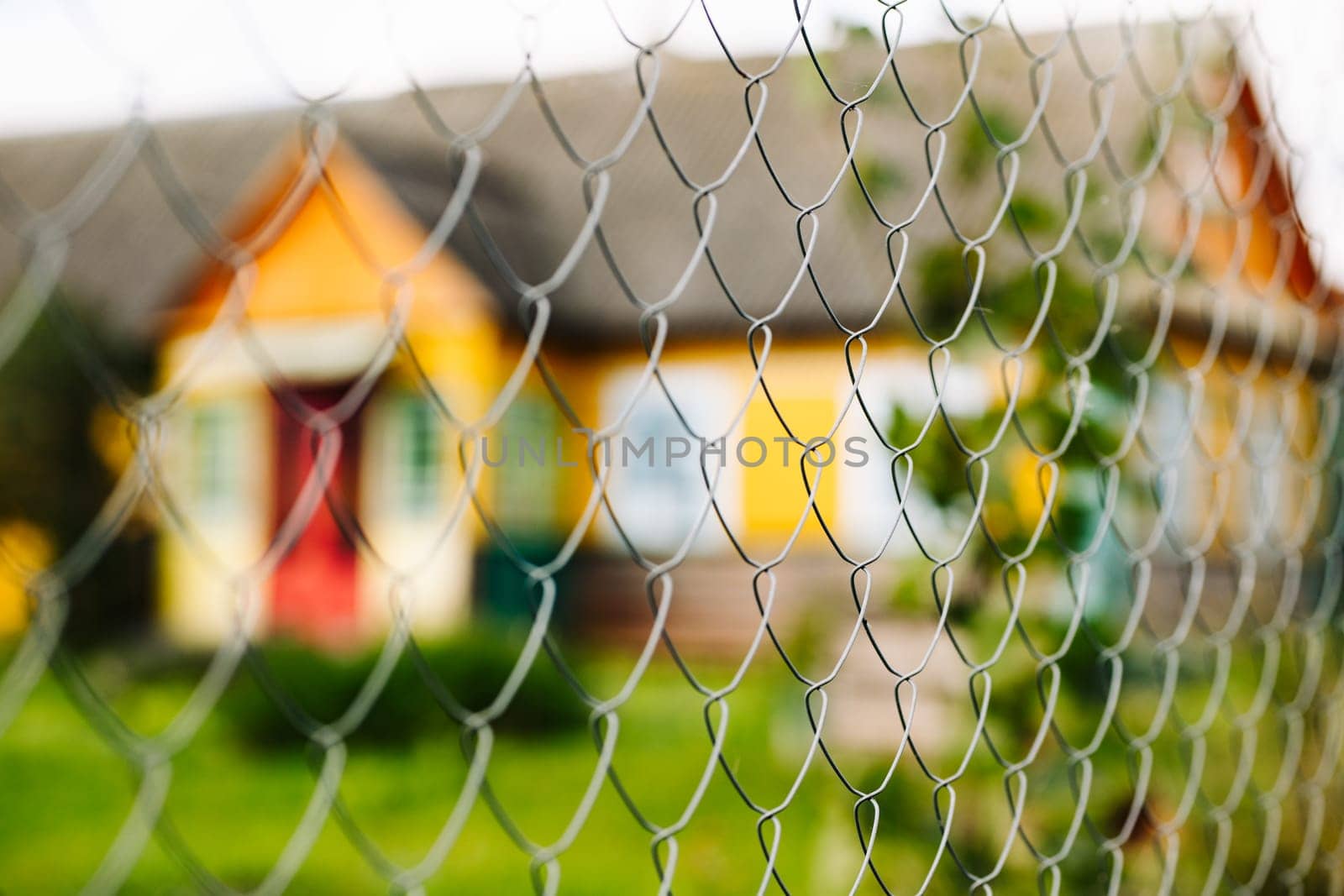 Metal mesh fence is separating viewer from colorful house with red door and green grass lawn