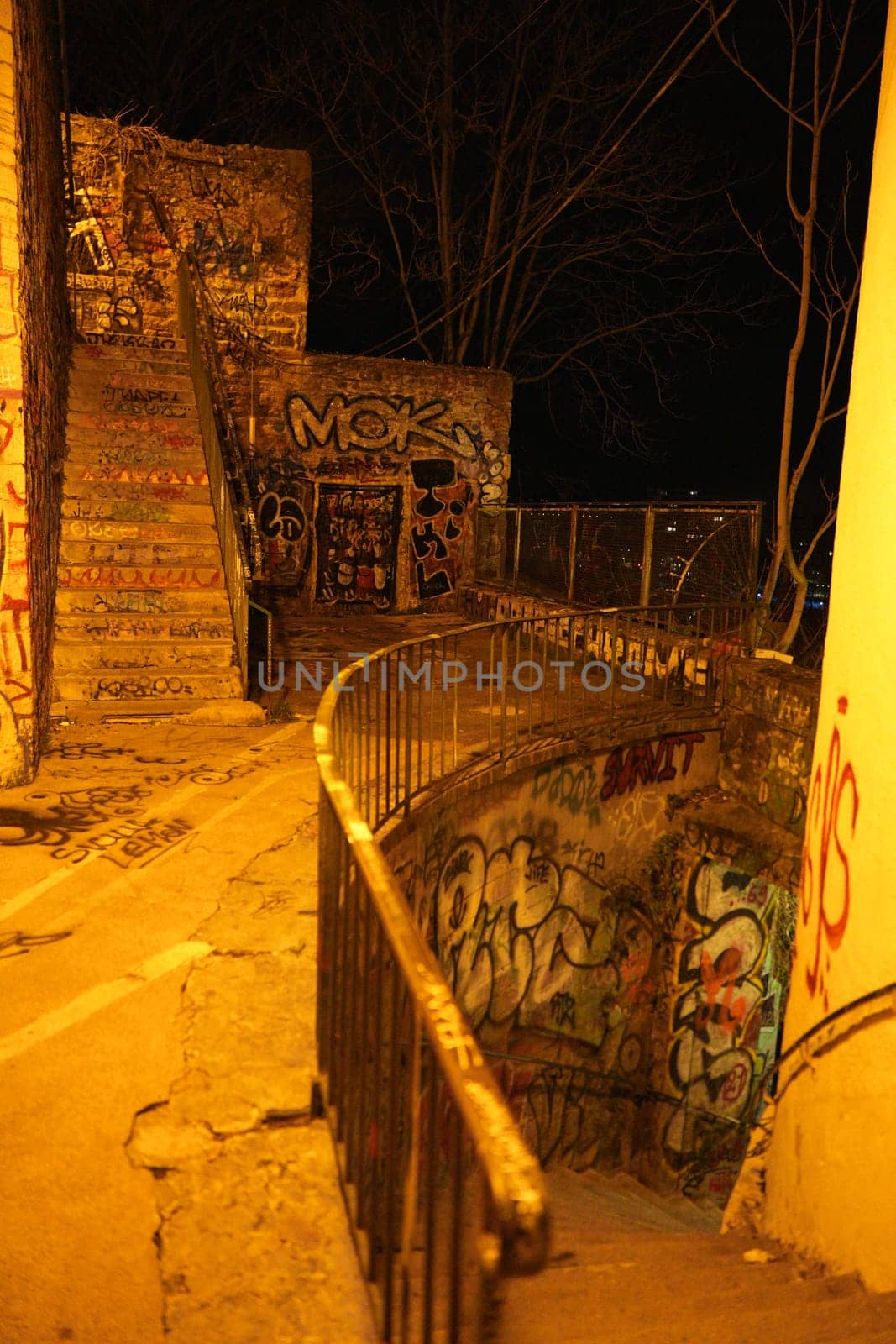 Nighttime view of an outdoor urban staircase spiraling downward, fully covered in colorful graffiti and dimly lit by orange streetlight.
