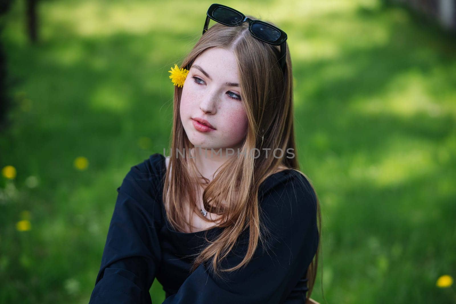 Portrait of a pensive teenage student with sunglasses and a yellow flower in her hair, enjoying a relaxing school day in a green park