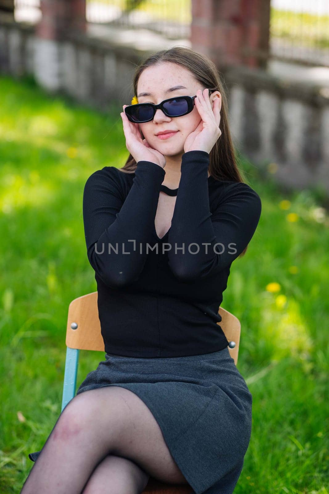 Female student taking a relaxing break outdoors, sitting on a chair in a sunny park, sporting sunglasses on a beautiful school day