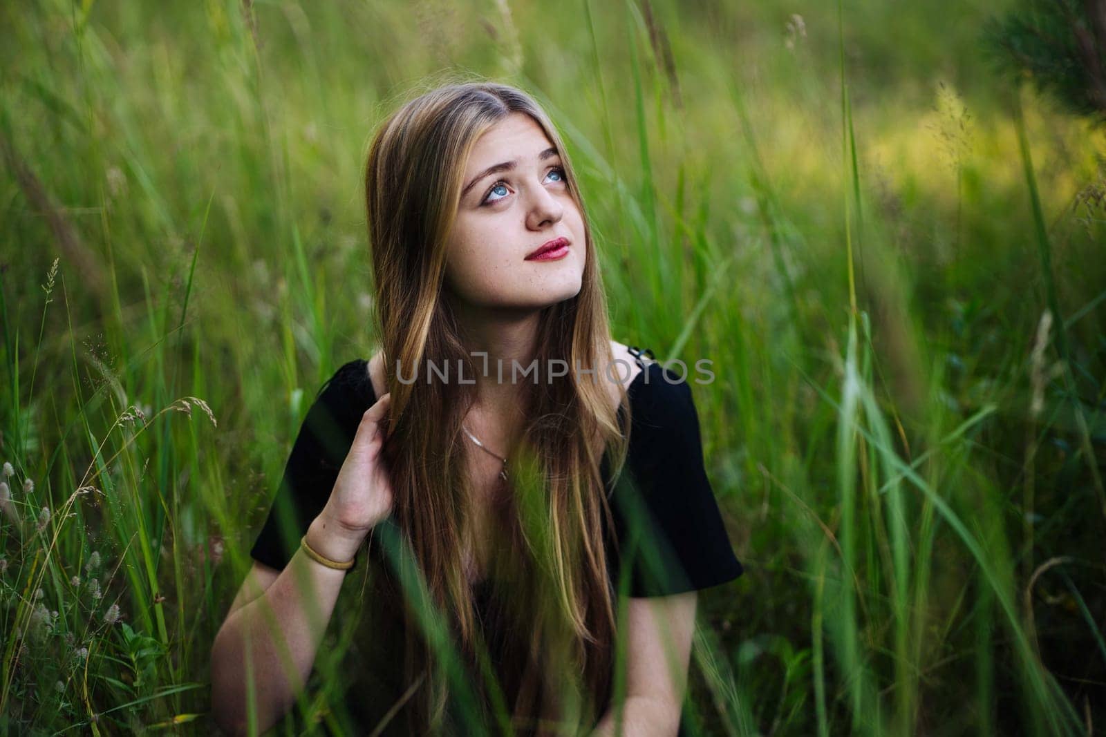 Young woman with long brown hair sitting in a field of tall grass, touching her hair and looking up, enjoying the peace and tranquility of nature