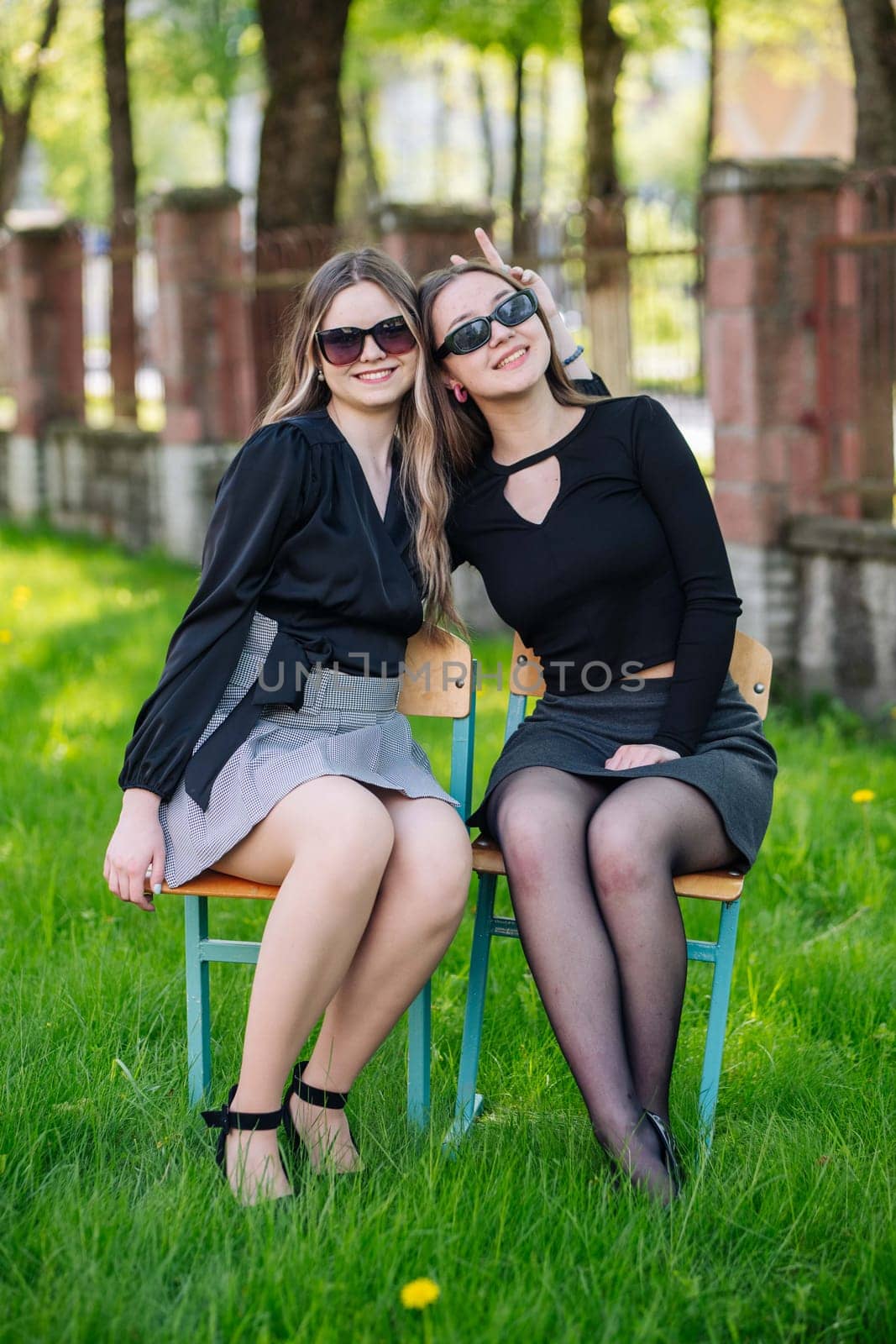 Two female students are enjoying their school day, sitting on chairs in the grassy schoolyard, smiling and making playful gestures