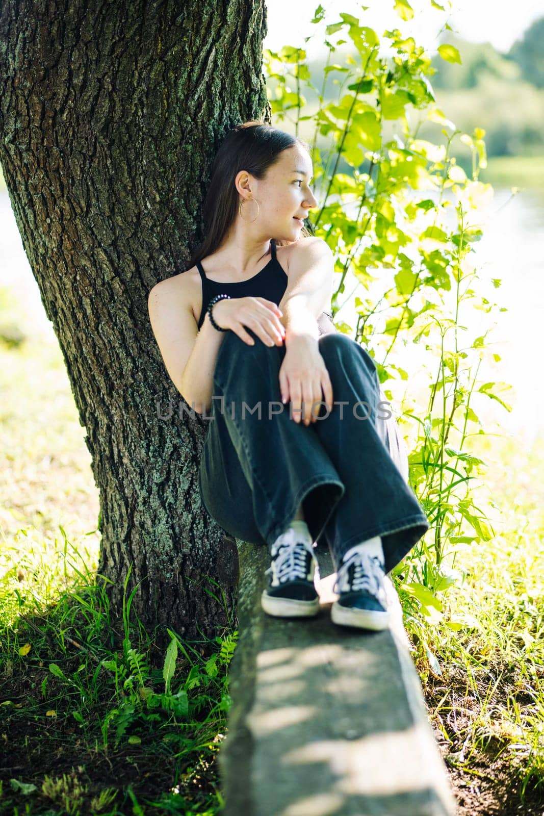 Serene female relaxing near lakeside tree, finding calm amid lush summer landscape, connecting with natural surroundings