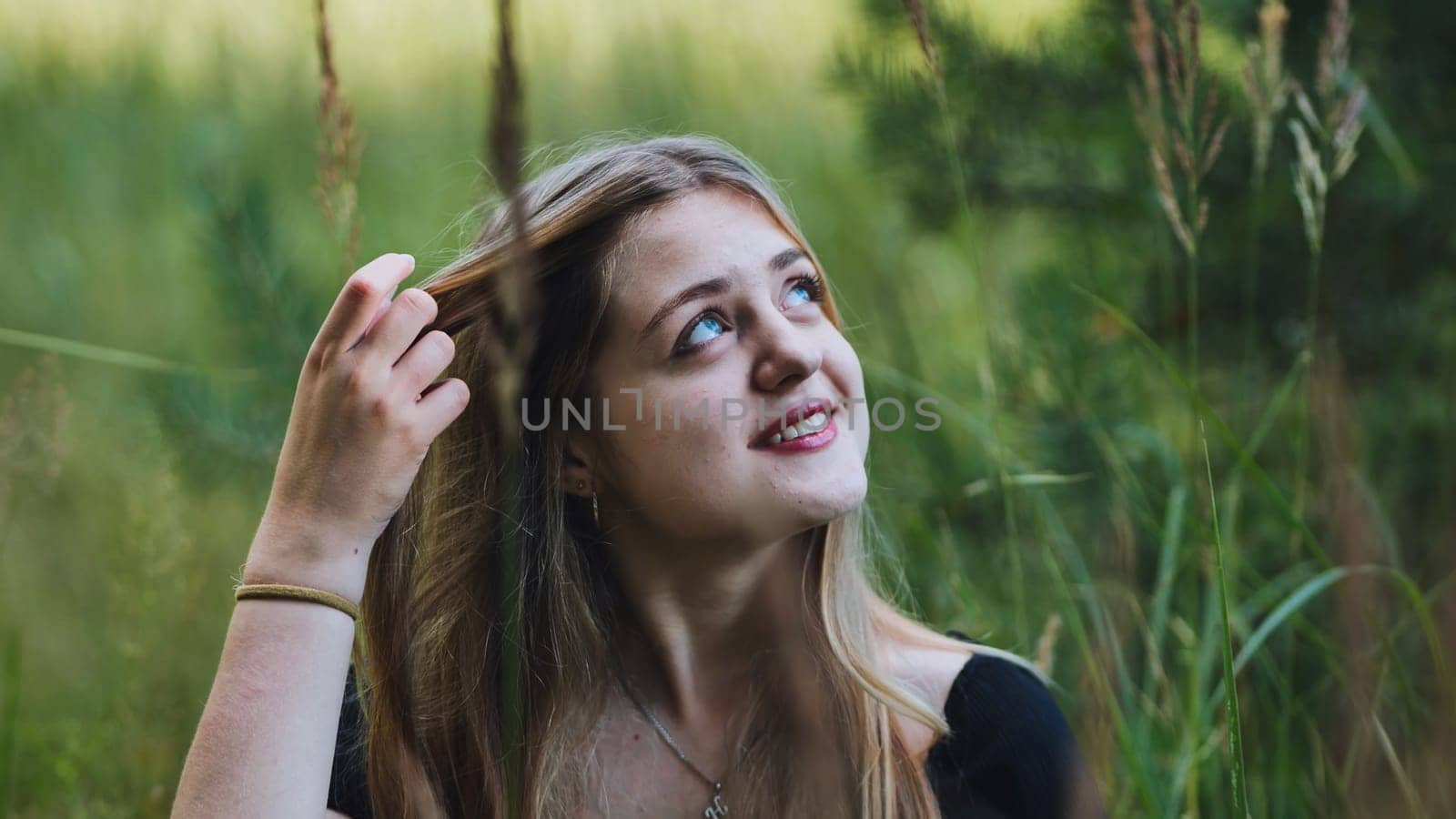 Blonde teenage student touching hair, gazing upward while basking in warm sunlight amid natural green surroundings