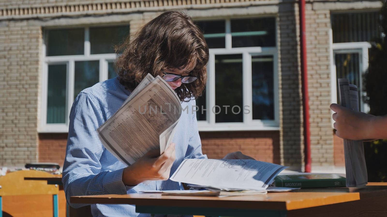 Teenage student reviewing test papers at an outdoor desk during the school day, focused on preparation under the warm sunlight