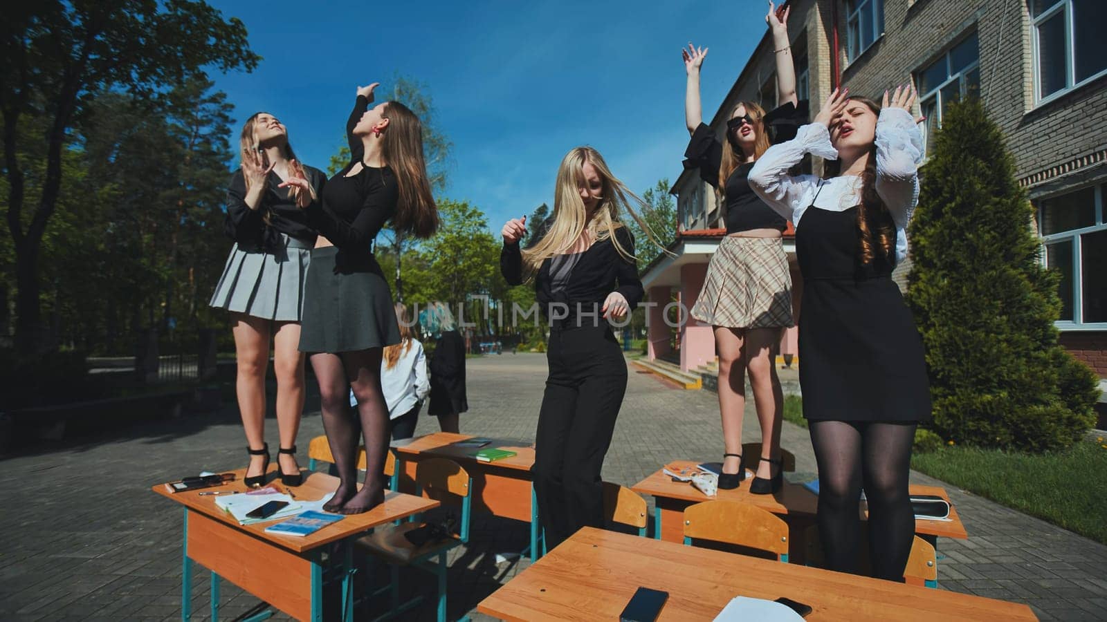 Five cheerful high school girls dancing on school desks in the schoolyard, celebrating the end of the school day
