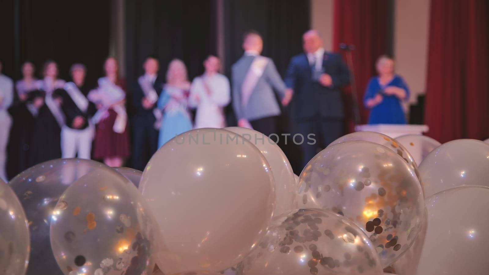 Filling the foreground, white and transparent balloons with gold and silver confetti contrast with blurred figures of graduating students and teachers on stage