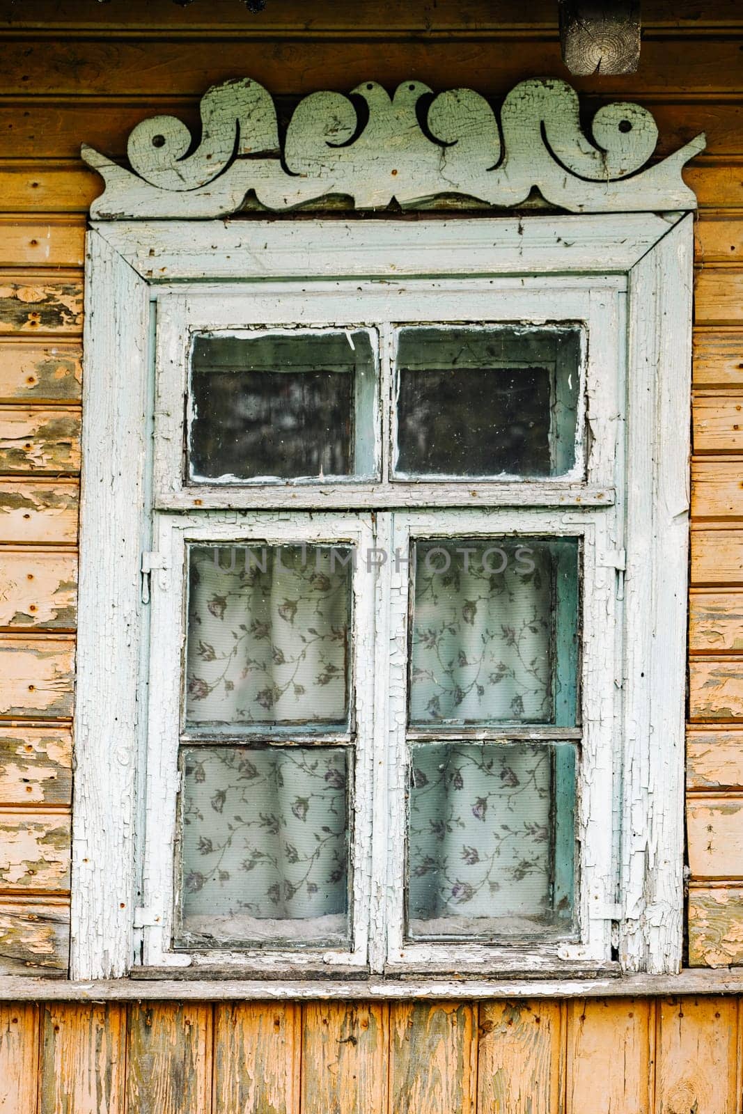 Weathered window with peeling paint, carved valance, and floral curtains, adding a touch of nostalgia to a traditional wooden house