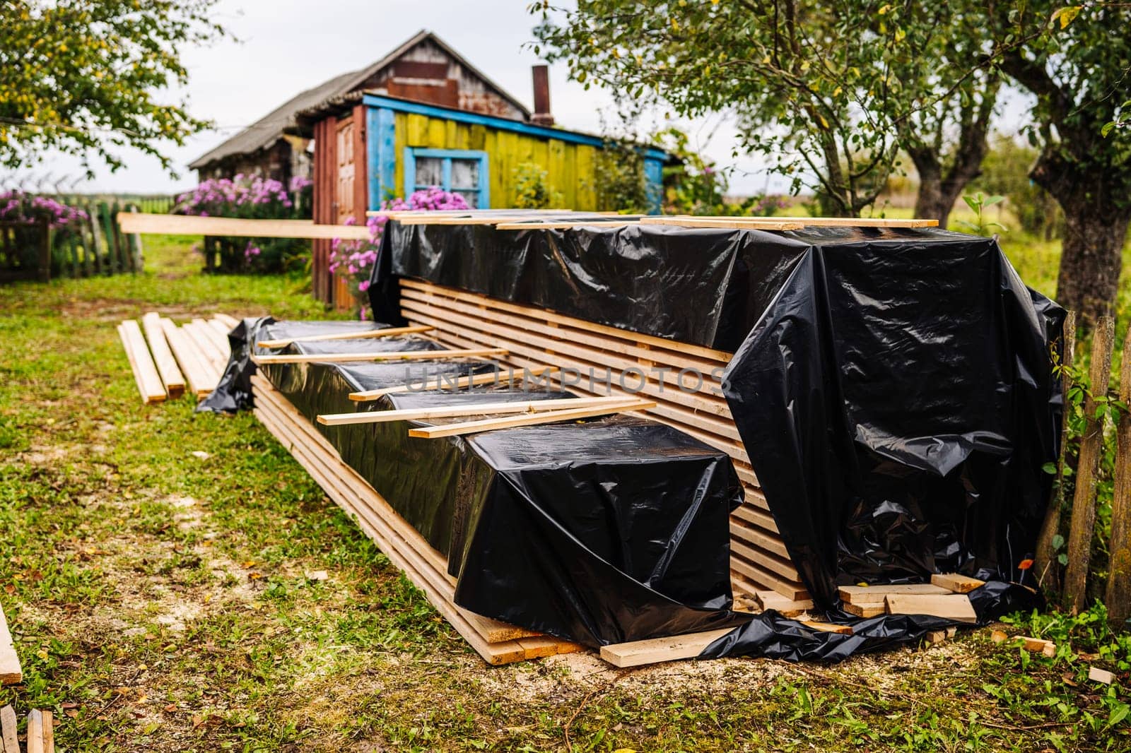Stacks of wooden planks covered with black plastic sheet material protecting from rain near colorful rural house in countryside