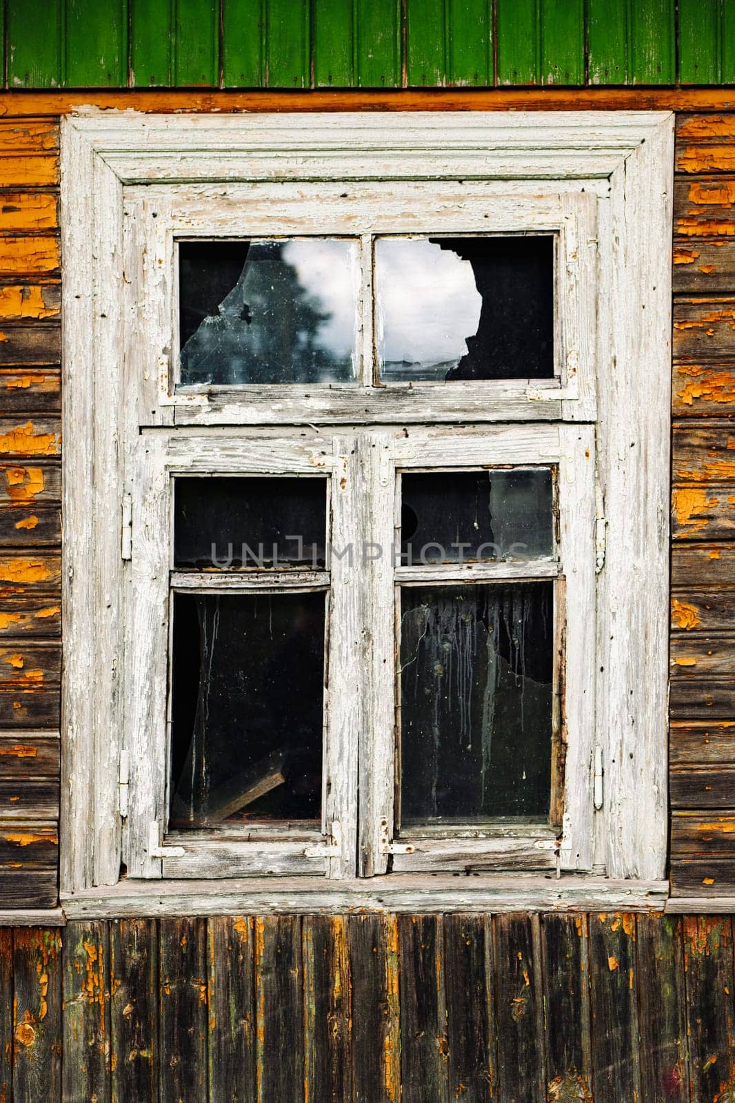 Broken window with peeling paint in old abandoned wooden house reflecting cloudy sky