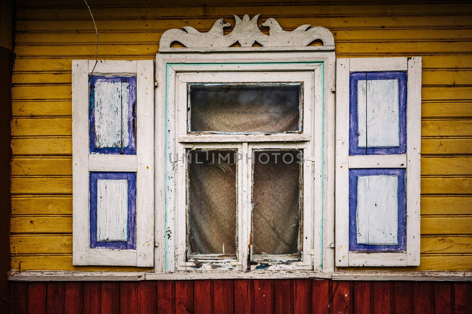 Weathered window with peeling paint and decorative shutters on a yellow wooden wall, suggesting age and decay