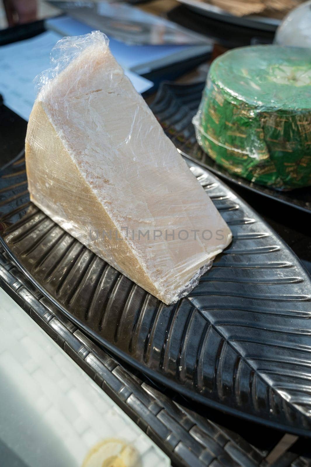 a piece of cheese in clear packaging at a farmers market. a large piece of cheese on a black oval plate.