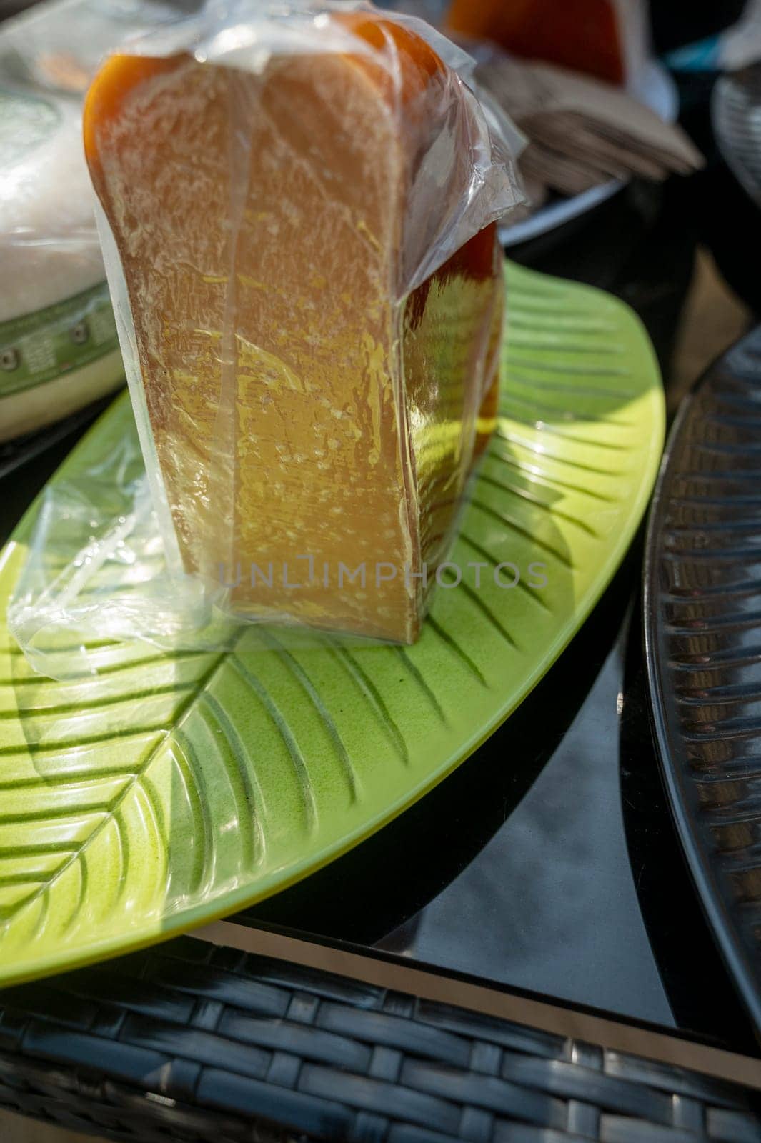 A piece of cheese, dark yellow in transparent packaging at a farmers market. Natural cheese at a city fair. A large piece of cheese on a green oval plate.