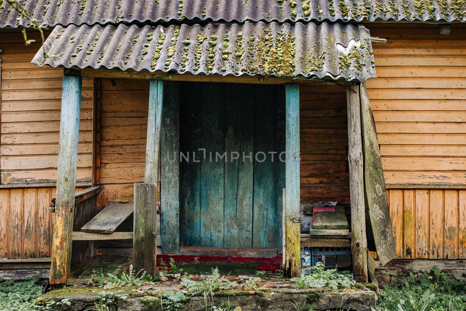 Porch of a decaying wooden house with moss and lichen growing on the roof and a weathered blue door