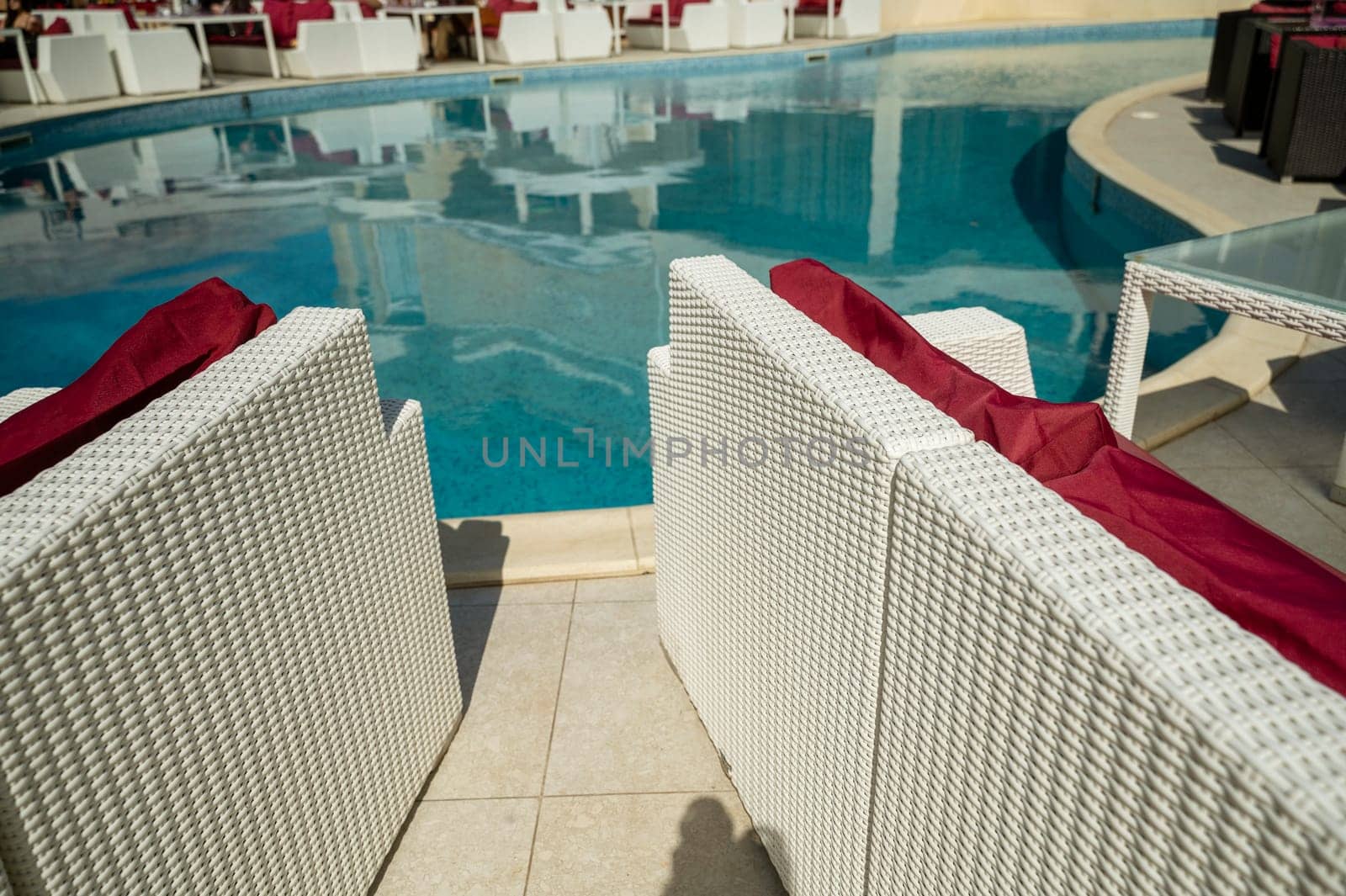 Bright blue swimming pool and dining area around it in the hotel. White wicker sofas with red pillows.