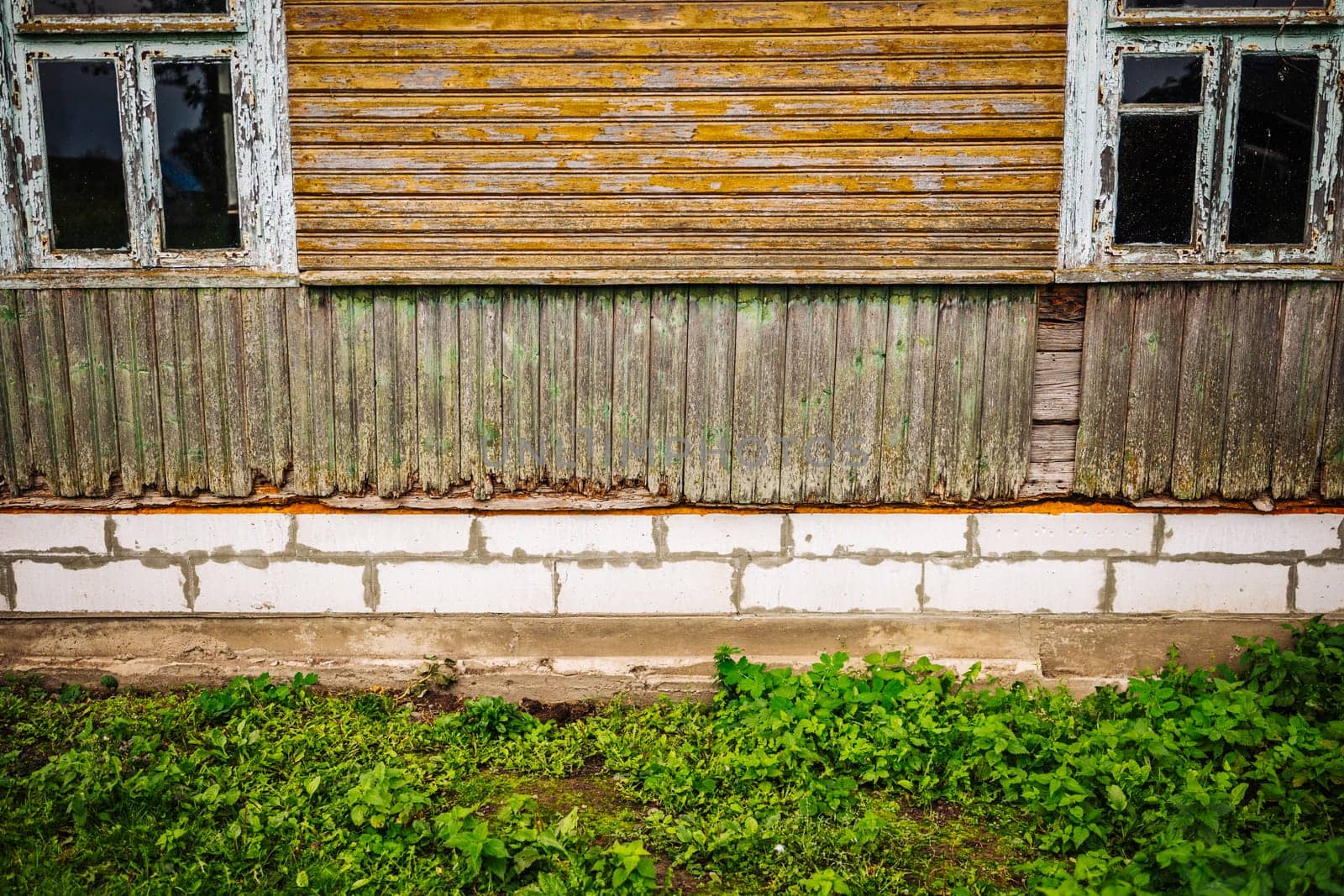 Weathered wooden wall with damaged planks and two windows showing foundation and grass