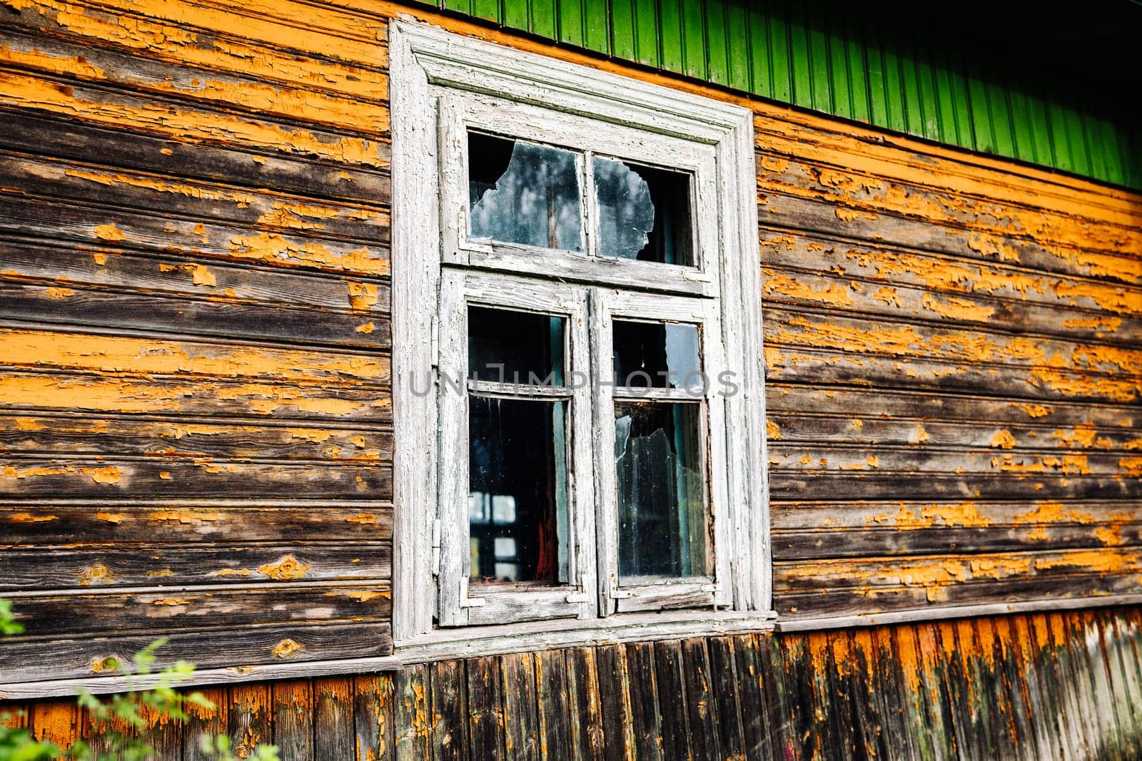 Old broken window in weathered wooden wall with peeling orange paint showing signs of decay