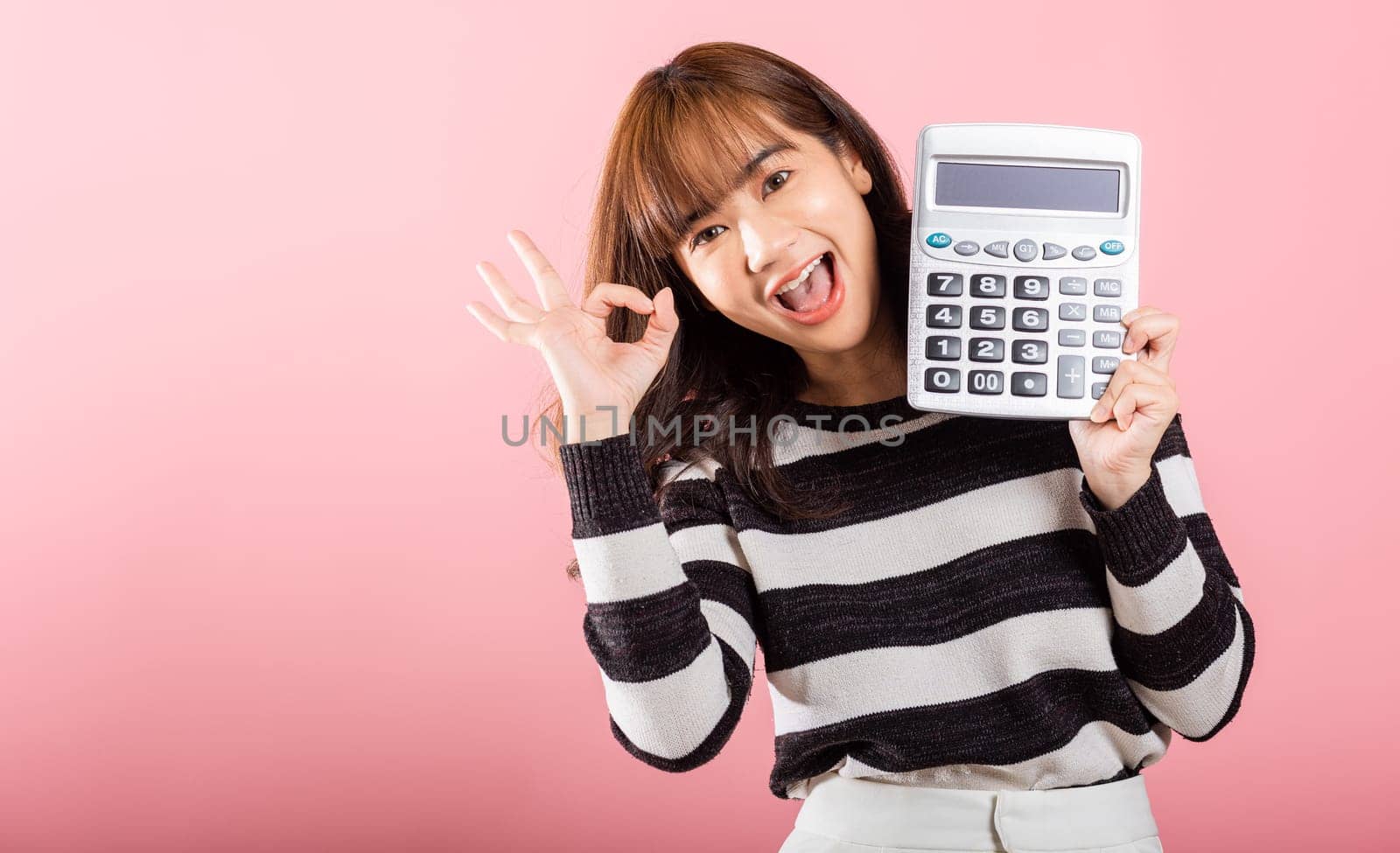 Tax day concept. Woman confident smiling holding electronic calculator and show OK gesture, excited happy Asian female studio shot isolated on pink background, Account and finance counting income