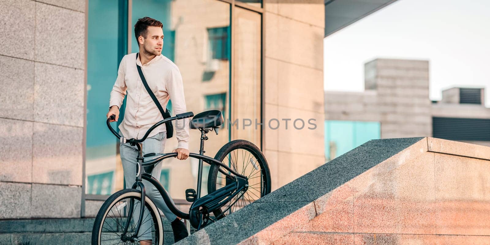 A young guy ascends steps with his bike in a modern urban environment. The warm light of sunset highlights his relaxed yet focused demeanor, showcasing a casual lifestyle choice, copy space