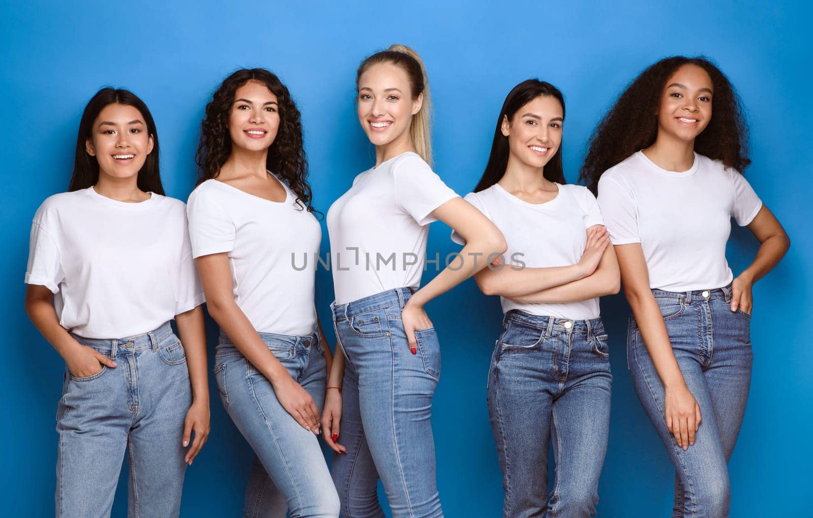 Pretty Multicultural Girls Smiling To Camera Standing On Blue Studio Background, Wearing Casual Jeans And T-Shirts. Diverse Beauty Concept.