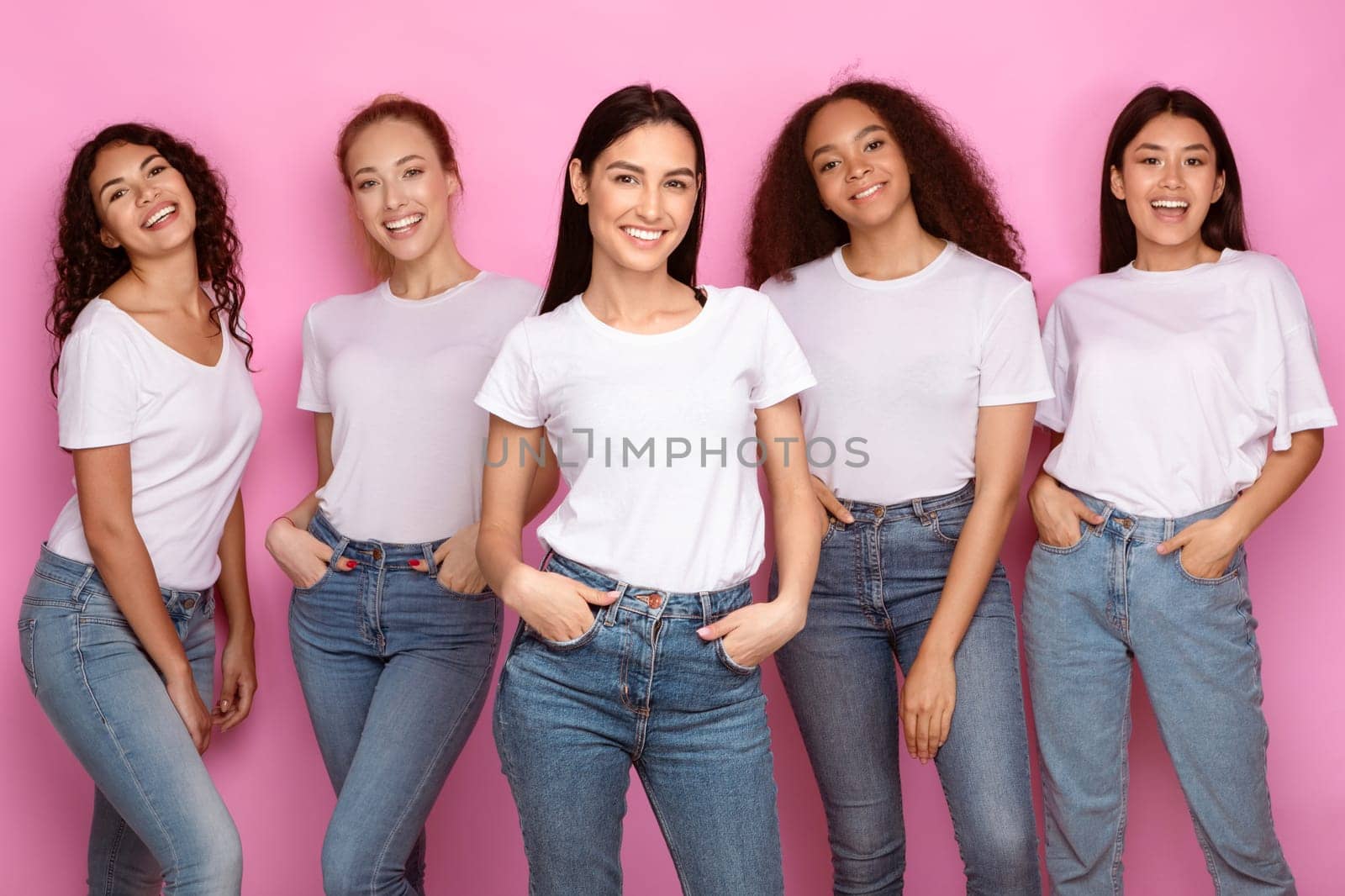 Five Pretty Mixed Young Ladies Standing And Smiling To Camera Posing On Pink Background In Studio, Wearing Casual Clothes T-Shirts And Jeans. Happy Females Group, Multiethnic Beauty Concept