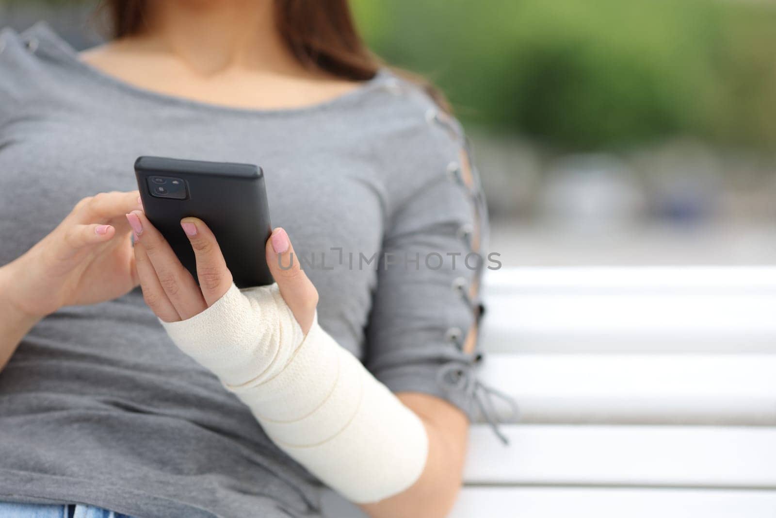 Close up portrait of a convalescent woman with bandaged arm using phone sitting on a bench in a park