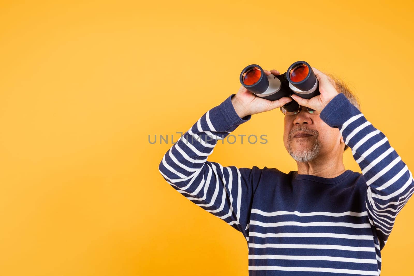 Portrait Asian smiling old man looking camera through binoculars studio shot isolated yellow background, senior man pensioner feeling surprised using binocular