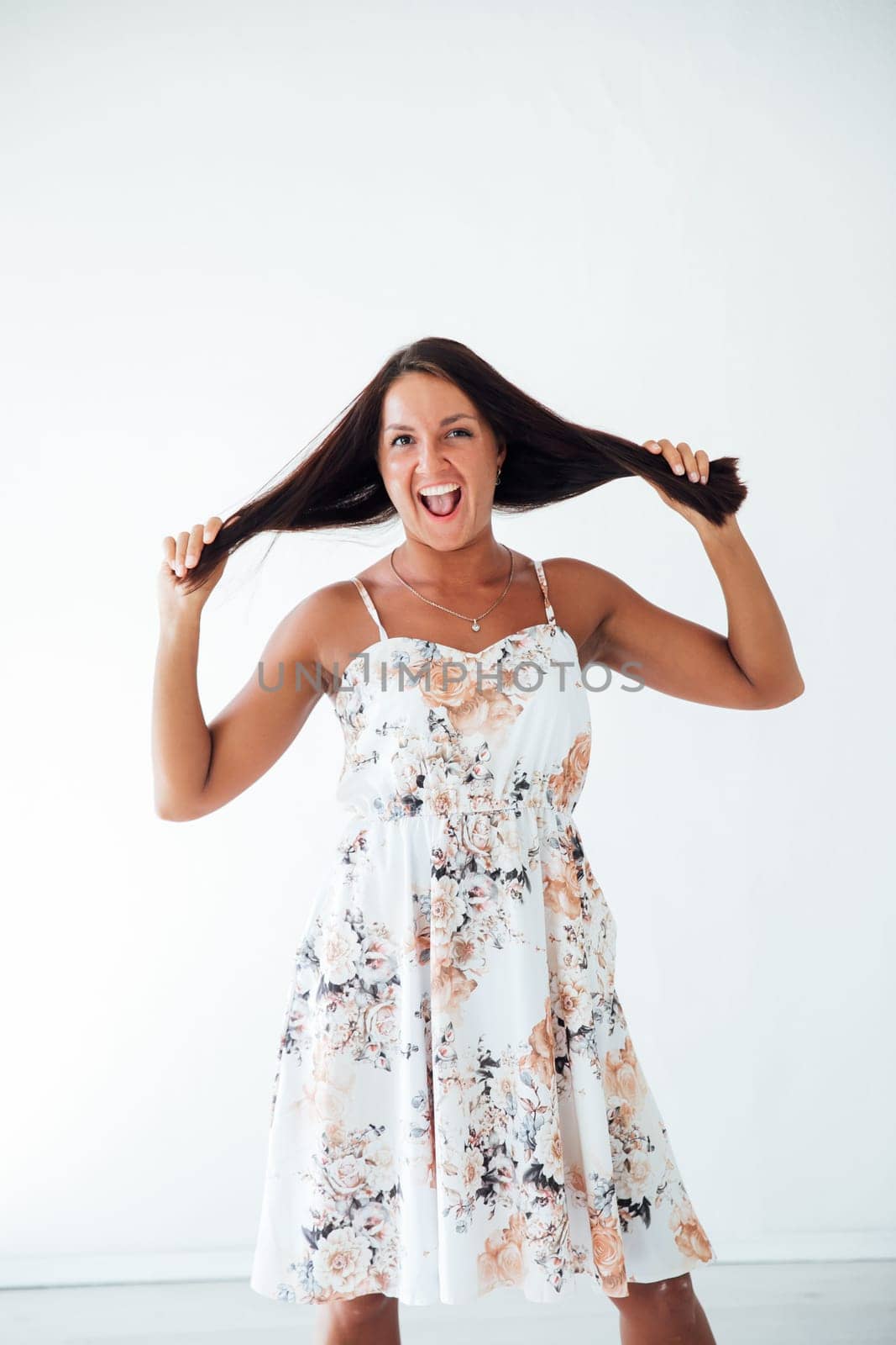 Brunette woman in summer dress holding her hair and laughing in white room