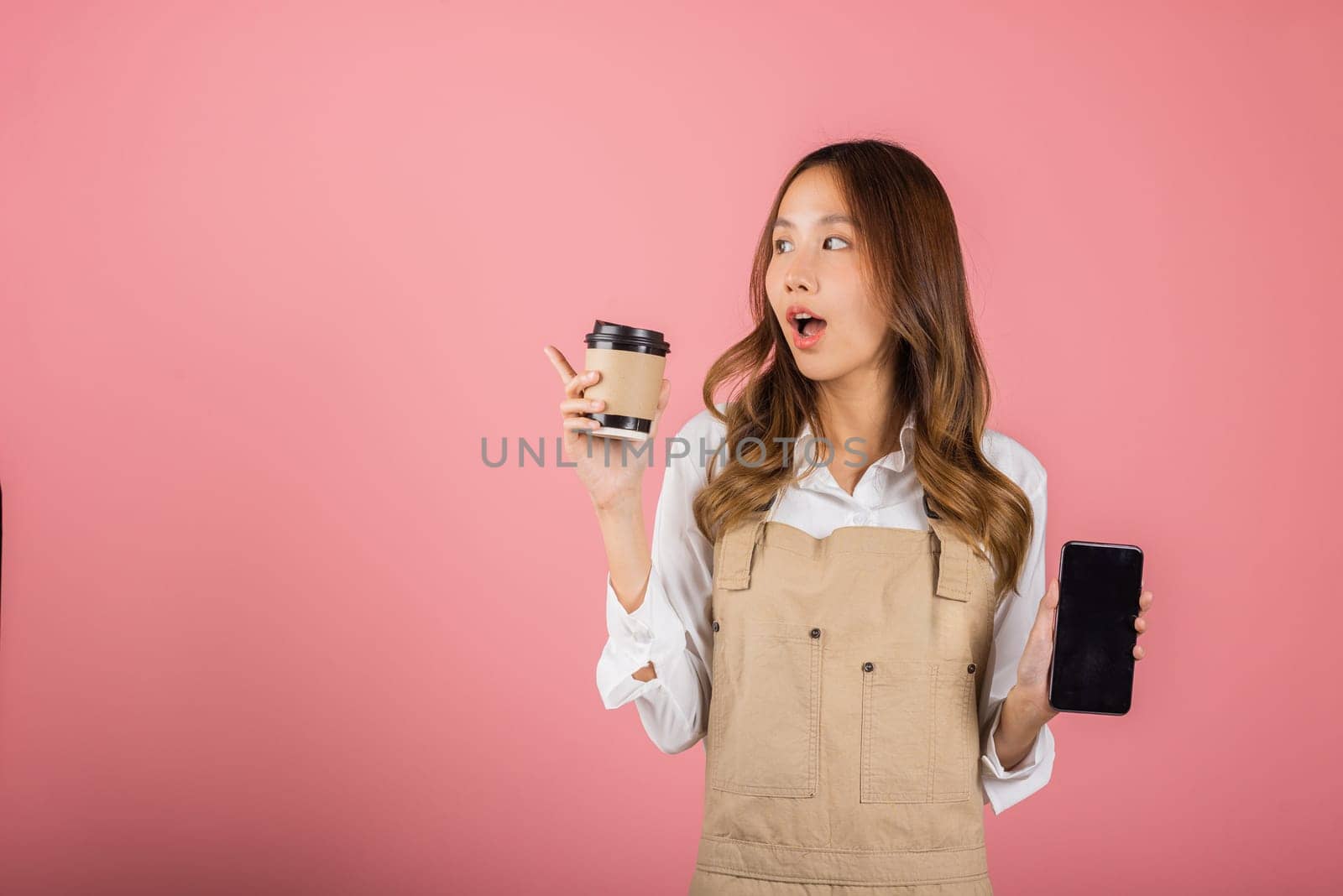Portrait Asian happy young woman barista bar tender coffee maker holding coffee tea hot cup and mobile phone studio shot isolated on pink background, female smiling hold takeaways cup and smartphone