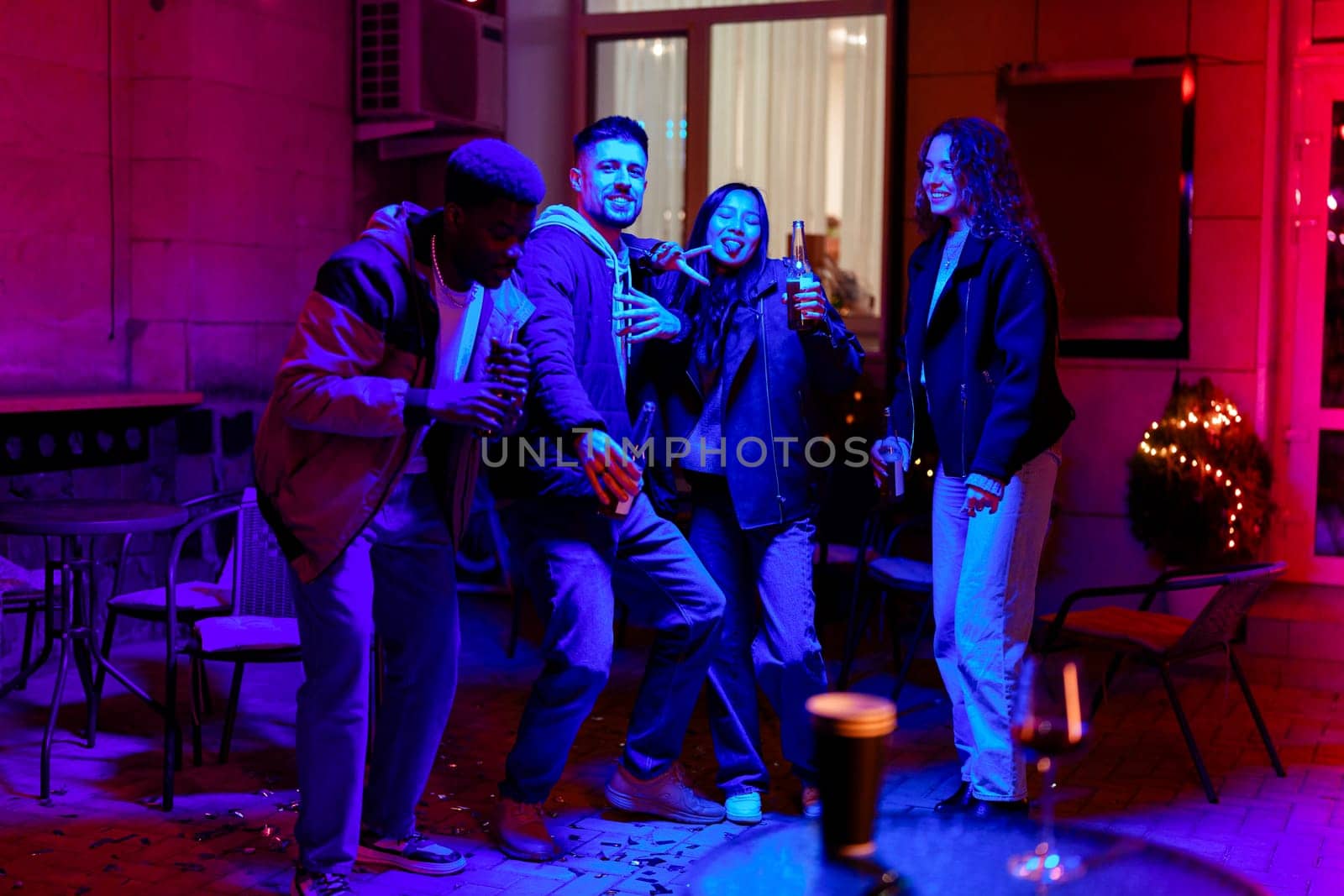 A group of friends are joyfully dancing together in a colorful outdoor space adorned with festive lights. They are celebrating and enjoying drinks in a cozy atmosphere.