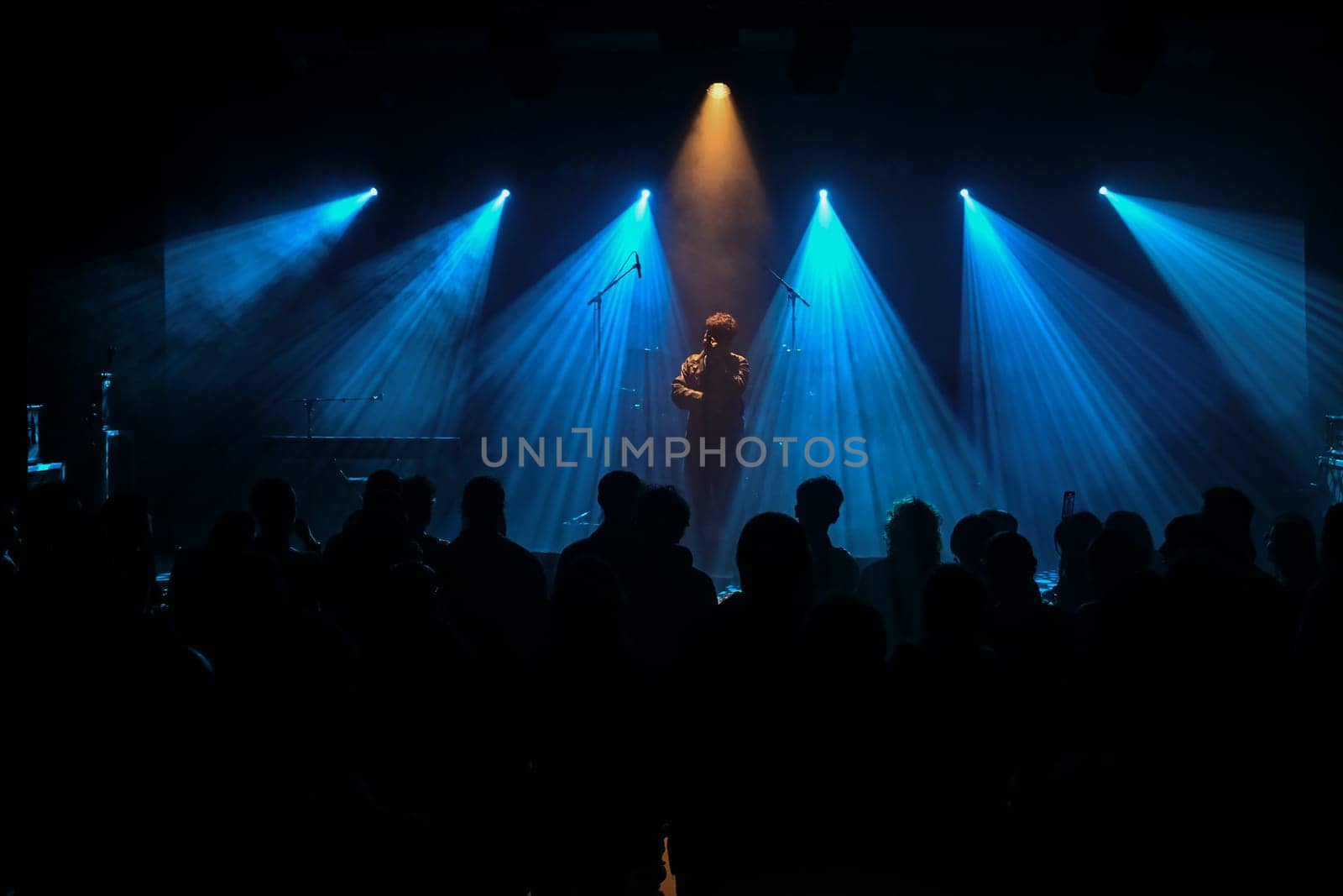 Musician stands center stage surrounded by blue lighting, creating a vibrant atmosphere. The audience watches intently, enjoying the live musical experience.
