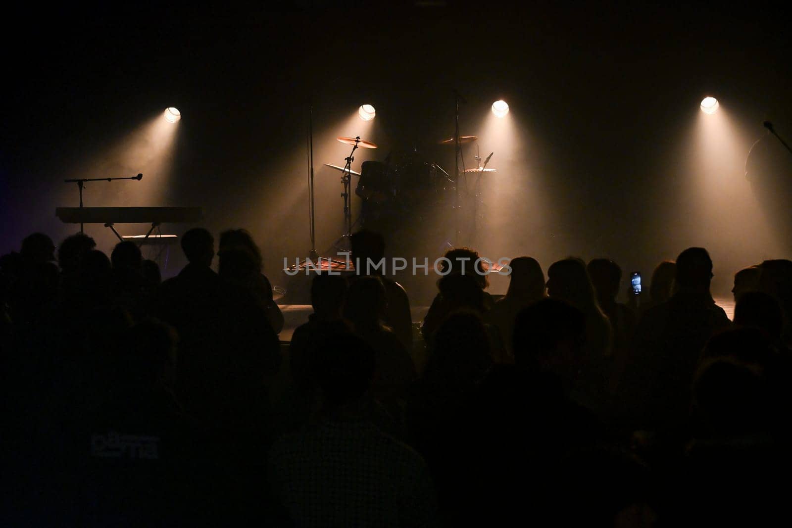 Audience gathers in a lively venue, eagerly anticipating the performance as stage lights illuminate the setup, creating a vibrant atmosphere filled with excitement and energy.