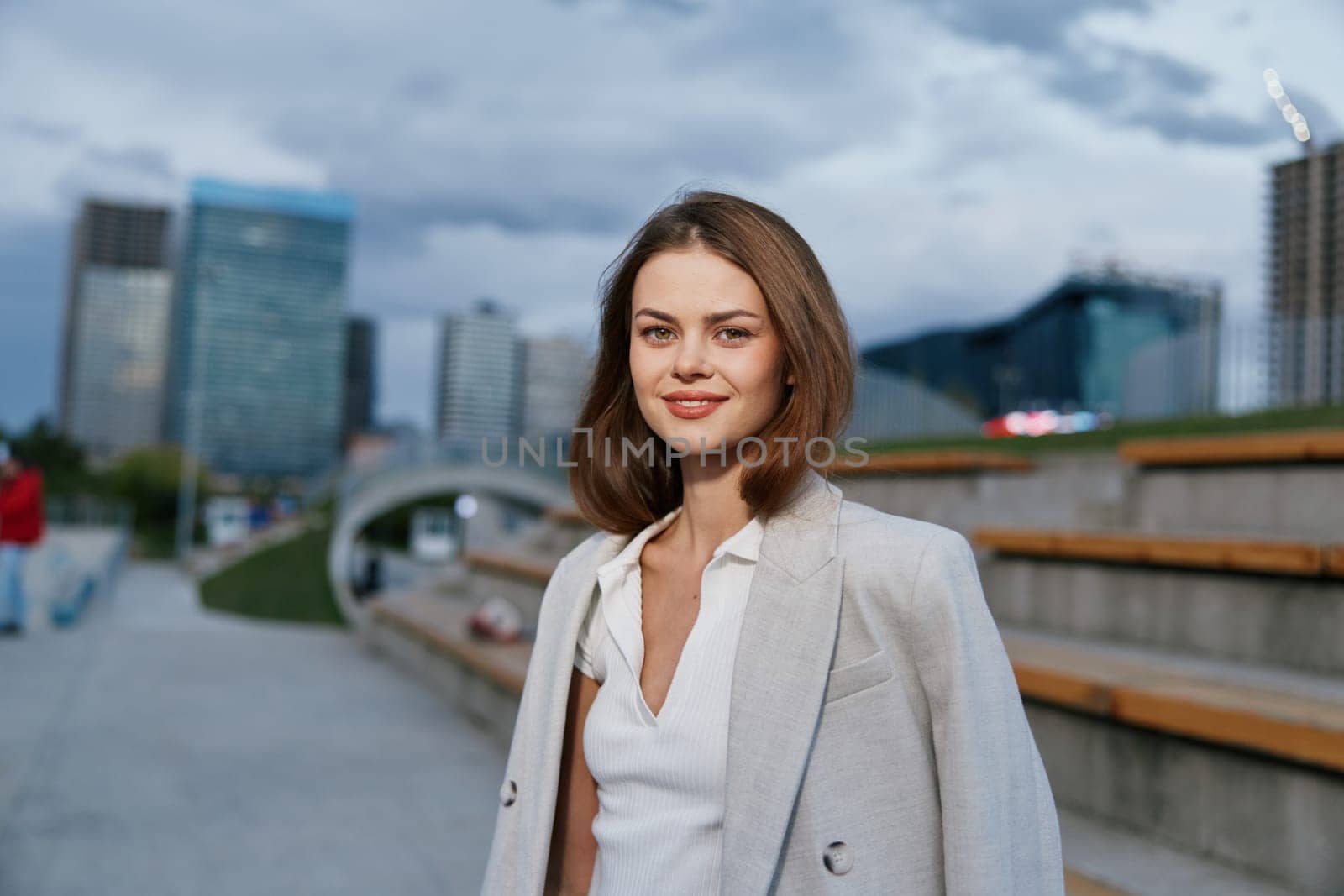 confident woman in business attire stands outdoors in urban environment with modern buildings and cloudy sky, showing casual style and professional appearance