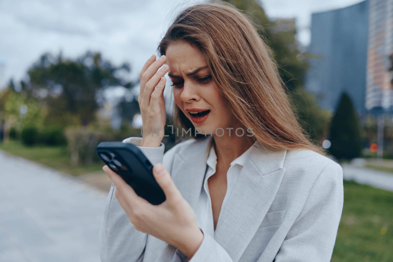 A distressed business woman in a light blazer looks at her smartphone with concern outdoors in an urban environment. Her emotion reflects the challenges of modern business.