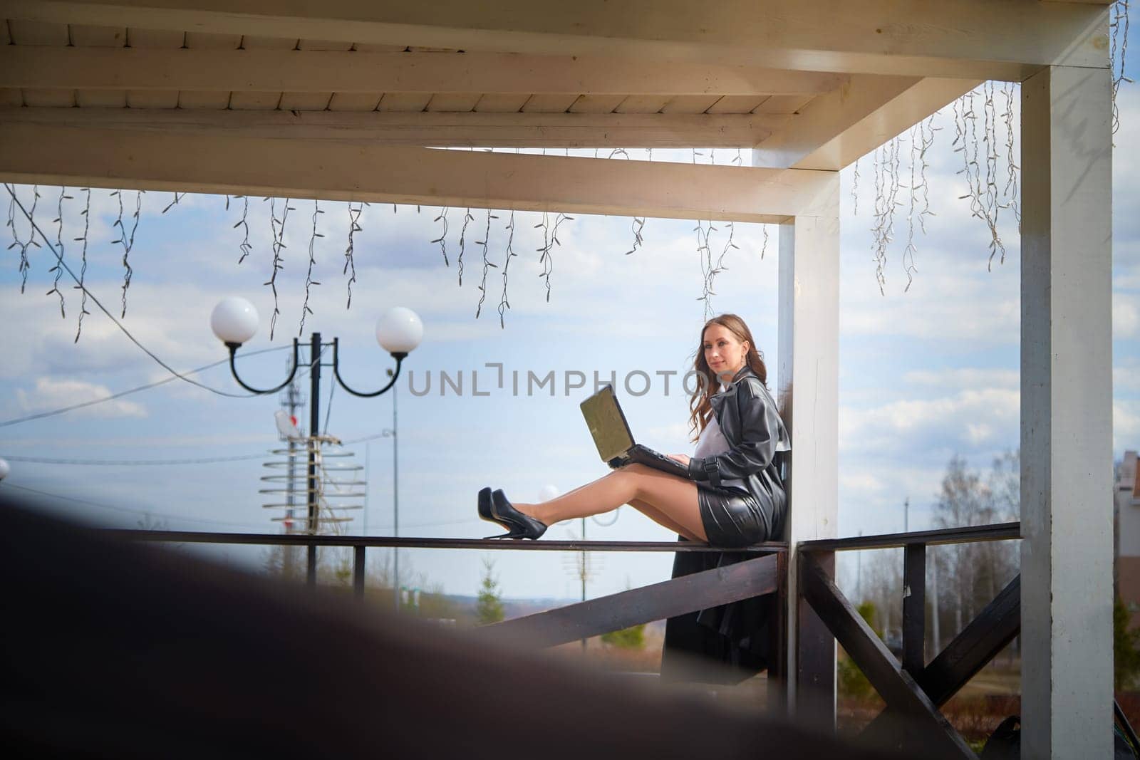 A woman focused on her laptop sits elegantly on a wooden railing in a sunny outdoor area with decorative lights.