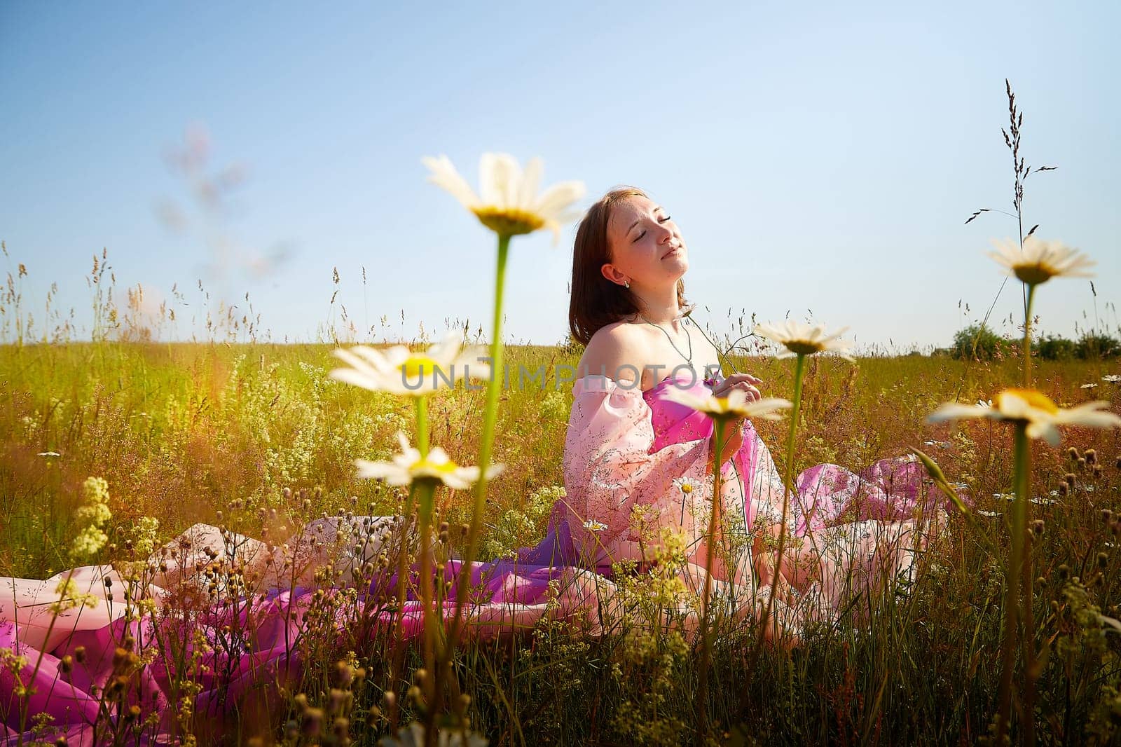 A woman wearing a vibrant dress sits peacefully in a flower-filled field, basking in the warmth of a sunny day, enjoying nature's beauty.