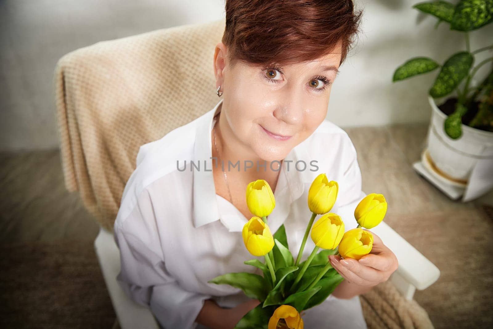 A smiling middle-aged woman holds a bouquet of tulips in her cozy room, celebrating Women's Day with joy.