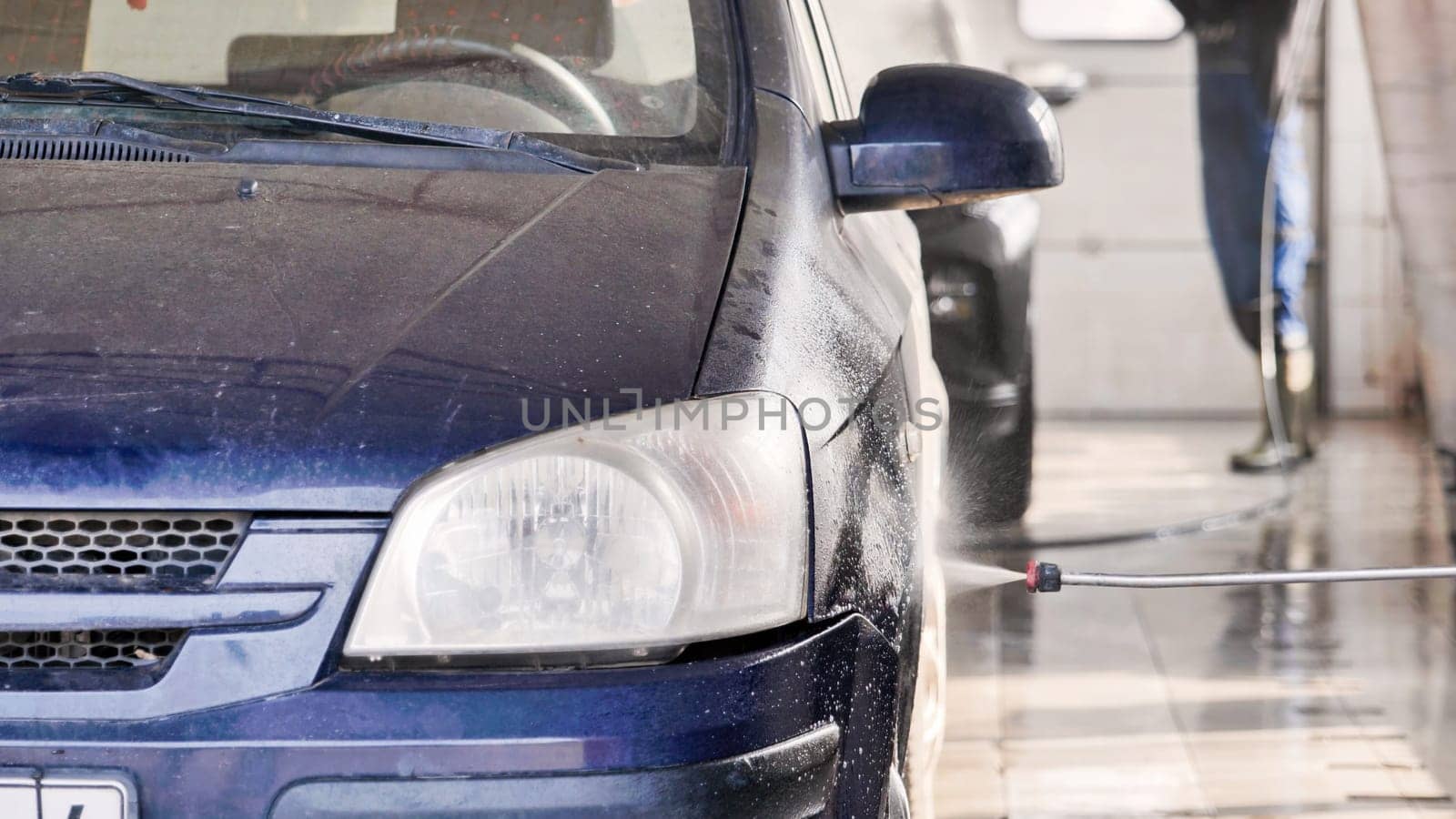 Car wash worker spraying water on dirty blue car using high pressure washer lance in service station