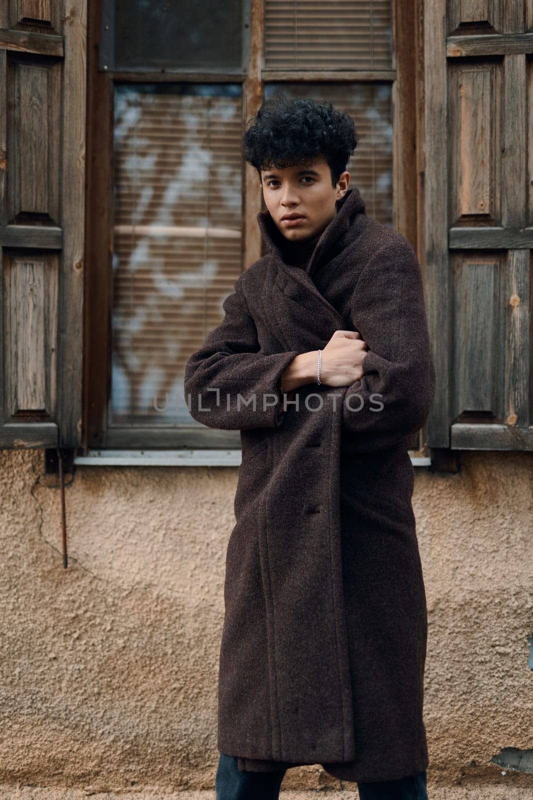 Fashionable young man in a brown coat posing in front of an old building, with hands on hips and looking at the camera.
