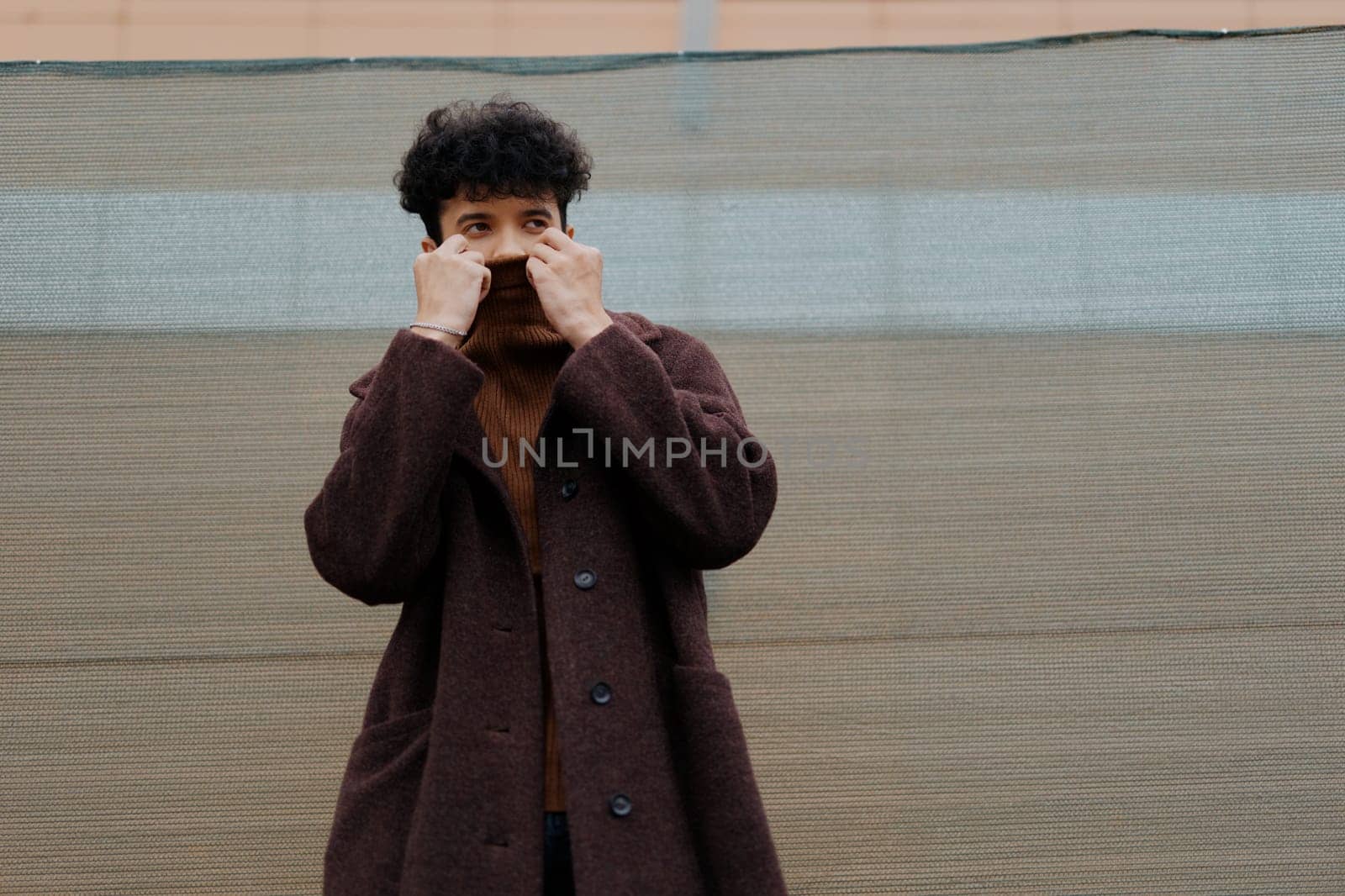 Fashion man in brown coat holding hands to face in front of green wall with brown tarp.