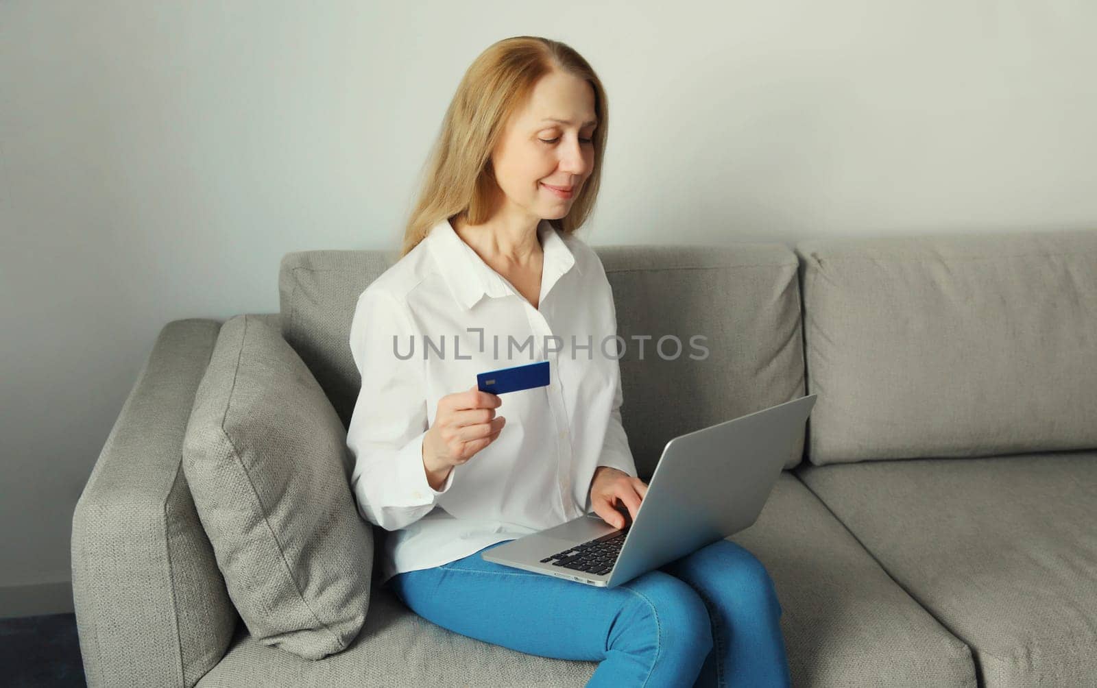 Happy smiling woman user holding plastic credit bank card, looking at laptop, shopping online or ordering goods via computer sitting on the couch in living room at home
